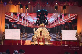A stage event photographed from the front. A man in saffron-red robes with a red head covering and multiple beaded necklaces sits on an ornate golden throne with a dark bronze multi-armed deity murti mounted above it. Two whiteboards flank the stage with state-of-consciousness diagrams. Tall trident staffs stand on the red-carpeted stage, and a large screen behind shows an arched palace hall with a checkered floor. Audience members are visible at the bottom edge in the foreground, partially cut off.