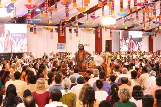 A large tented hall filled with seated devotees facing a raised dais. A man with long dark hair and a full beard stands at centre, wearing an orange-saffron garment and a dark patterned shawl, one hand on his hip, beside a gold-and-cushioned seat. Two large screens on either side show close-up images of a similar bearded figure. Overhead hang rows of colourful decorative lamps with tassels and oil-lamp style fixtures. Many people sit cross-legged in white, cream, yellow and orange clothing, while others sit in chairs at the sides.
