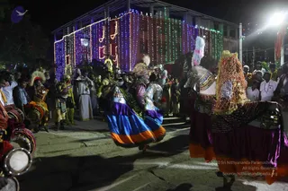 Night procession with folk dancers in elaborate costumes in front of a building decorated with colourful string lights and illuminated letters. In the centre, a dancer in a blue, purple and orange skirt with a gold-and-red headdress and a silver-patterned bodice leaps forward. To the right, a drummer wears a tall plume of white and pink feathers and a sequined, mesh-patterned purple costume. Behind them, a crowd of onlookers and devotees in white shirts and dhotis watches. Strong white floodlight illuminates the scene from the upper right.