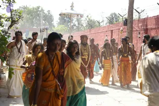 A procession moves along a paved path beside a red-painted boundary wall topped with barbed wire, under a bright sky. Around him, devotees wear saffron and orange saris and dhotis, yellow and green garments, and rudraksha malas, with some carrying marigold garlands. Men in white dhotis and shawls walk nearby, one holding a small decorative staff. An ornate saffron-and-gold parasol rises above the group, and a trident-tipped staff appears at the upper left. Dust hangs in the air near the foliage at left.