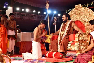 A stage event under a tent with stage lights and a metal truss overhead. A seated man in a rust-brown robe with a long swept-back hair, beard, a long white flower garland and gold ornaments sits on an ornate gold throne with red cushions, a person by his long swept-back hair, ornate gold throne-style seat and ceremonial gold necklaces. A shirtless young boy in a white dhoti kneels before him, bowing with folded hands and holding a yellow cloth or envelope. Behind the boy, a saffron-robed attendant and a bare-chested man in white dhoti stand. A golden trident stands to the side, and a woman in a maroon sari sits at the right edge.