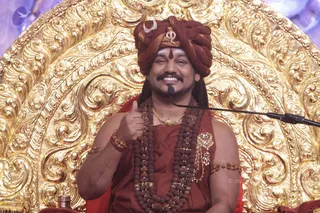 A smiling man seated on an ornate gilded throne with elaborate repoussé scrollwork behind him. He wears a maroon-red turban with a crescent moon and trident emblem, a red-brown robe, long rudraksha mala necklaces, a gold pendant, gold armlets and a red thread bracelet. He gives a thumbs-up gesture and sits before a microphone on a stand. Swamiji (probable): the appearance cues are a maroon turban, rudraksha malas, ornate gold jewellery and a throne setting, consistent with the page's reference to a discourse.