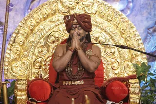 A photograph of a seated man in a rust-red turban and matching rust-red robes, hands pressed together in anjali mudra at chest level. He wears layered rudraksha mala necklaces, gold bangles, a gold armband, a jewelled gold waist belt and a small gold pendant. He sits on an ornate gold throne with carved scrollwork and red cushions, with a microphone on a stand beside him and green foliage at the right edge.