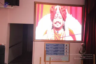 A photograph of a wall-mounted screen in a hall with pink walls and red curtains. On the screen, a smiling man wearing an orange-red turban with a tilak and a white gem on the forehead, a saffron shawl, and a long beaded necklace speaks into a microphone; the page indicates he is Swamiji, though the appearance cues are only partly distinctive. Below the screen hangs a framed sign in Tamil and English prohibiting photography, video camera, mobile camera and audio recording, marked 'Strictly Prohibited'. A trident-topped brass finial stands at the right near red drapes, and a mic cable runs across the frame.