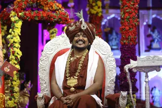 A smiling man sits on an ornate silver-and-white throne with carved lion-head armrests, hands folded in his lap. He wears a deep maroon turban with a silver ornament and a white feather, a maroon robe, a white shawl draped over the shoulders, and a long gold ceremonial necklace with a large gold pendant. Behind him are arches of red and yellow flower garlands and purple-lit pillars, with a small deity murti visible on the right. A silver lectern stands at the right edge. The man is one person; he is the appearance-based candidate, showing a turban, ornate gold necklaces and saffron-toned robes, consistent with the page's Swamiji reference, and is labeled Swamiji (probable).
