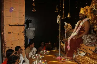 A man seated on an ornate gold throne draped with a leopard-print and tiger-striped cloth, wearing a gold crown, a pink and white flower garland, and a rust-red dhoti-style garment, with bare feet. He smiles toward a small group of women seated on the floor at left, one in a white sari with a rudraksha mala, another in white with gold jewelry. Silver plates hold flowers and offerings on a low golden table in the foreground, and a silver standing staff with a white cord stands beside the throne. Brass oil lamps and bells hang from the ceiling above, and a video camera on a tripod is at left. The setting is a dark room with string lights. The man has long swept-back hair and a crown, but the visible cues are limited to crown, garland and throne, so the identity is uncertain.