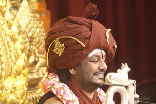 A smiling man in a rust-brown turban adorned with gold ornaments and a gold jewelled brooch sits beside a white marble Nandi figure. He wears a garland of pink flowers and white beads, a rust-brown shawl, and a white tilak on his forehead. Behind him stands ornate gold-leaf carved decoration against a deep red curtain. a person: long-haired presentation with a turban, gold ornaments, rudraksha-free but ceremonial garland and ornate clothing, seated as a speaker at the event.