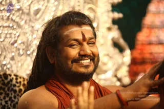 A smiling bearded man with long dark hair and a red-and-white tilak on his forehead sits in front of an ornate silver-embossed backdrop, with a leopard-print cloth at the lower left. He wears an orange-red wrap over bare shoulders and a red thread bracelet on his wrist, and his right hand is raised in a blessing gesture toward the camera. A carved ornamental structure in warm orange tones appears at the right edge. a person appears by his long hair, orange wrap and ceremonial presentation, but the appearance is only partly clear, so identification is uncertain.
