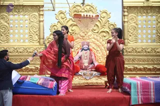 A stage performance with a golden ornate backdrop and a golden hooded cobra canopy above a large seated figure. A woman in a red and pink sari with long dark hair stands at center holding a microphone toward a man in a dark suit at left, who holds a clipboard. Two women in rust-brown and orange-red clothing stand at right and behind, gesturing with raised palms. Seated behind them on a golden throne with red cushions and a tiger-skin cloth is a large printed or painted depiction of a figure with an orange turban, orange robes, rudraksha-style beads and a gold necklace in meditation pose. A red carpet covers the stage and striped cloths drape the platforms.