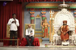 A stage event with a standing man in a white shirt and camouflage trousers holding a microphone and speaking. Beside him a bearded older man in a white shirt and dark trousers sits on a gold-trimmed red-cushioned chair. On the right, a seated figure in saffron-orange robes, a saffron turban, rudraksha malas and gold armbands sits on an ornate silver throne with leopard-print cushions; the appearance is consistent with a probable Swamiji (probable) in person. Behind them stands a painted deity murti with four arms holding a trident, drum and other objects, and a silver trident stands near the throne. Red velvet curtains and painted columns frame the stage.