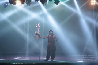 A man stands alone on a dimly lit stage under stage spotlights and haze, holding a tall silver trident (trishula) upright in his left hand with a small damaru drum hanging from it. He wears a dark maroon-red dhoti and matching upper cloth, has long dark hair, a tilak on his forehead and a rudraksha-style mala, and rests his other hand on his hip. The beams of light fan out across the background as he poses on the platform.