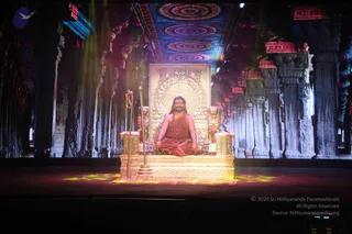 A man with long dark hair and a beard sits cross-legged on an ornate gold throne on a stage. He wears a saffron-orange robe, a long rudraksha mala and a gold necklace. A golden trident stands beside the throne and a golden staff-like rod stands on its other side. Behind him, a large projected backdrop shows a carved stone temple hall with pillars, a patterned ceiling of concentric circles and red-and-gold hanging decorations. Golden lion figures flank the throne and the stage floor has glowing yellow light patterns. Swamiji (probable): the seated figure has long swept-back hair, saffron robe, rudraksha mala and gold necklace, consistent with the page's reference to Swamiji.