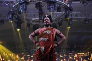 A photograph of a man standing on a stage under a truss rig of stage lights with yellow beams. He wears a rust-red draped garment with a leopard-print patterned sash across the chest and waist, red beaded armbands and a red-beaded necklace, a gold tilak and a gold ornament in his hair. His hands rest on his hips as he gazes upward. The stage floor shows a warm illuminated screen at the bottom edge.