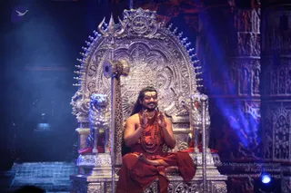 A photograph of a man seated cross-legged on an ornate silver throne on a dimly lit stage with blue light. He wears an orange-saffron robe, a long rudraksha mala with multiple strands, and a gold chain around the neck. His hands are raised in a greeting gesture, and he has long hair swept back. Two silver lion figures flank the throne, a silver trident and a leopard-print covered staff stand beside it, and a carved silver arch with a central emblem rises behind him. The background shows large carved temple-style pillars. Swamiji (probable): the orange robes, long hair and rudraksha malas match the page's description of him at this event, and he is seated on a throne with lion figures in saffron dress. Only one person is physically present.