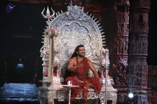 A seated man in an ornate silver throne with a trident and a damaru drum mounted on its back, flanked by carved lion figures. He wears a saffron-orange robe with a bare upper body, long dark hair swept back, a rudraksha mala, and a gold necklace, and he gestures with his right hand while smiling. A small white cup sits on a side table beside him. Behind the throne rise carved red-stone temple pillars, and a microphone stands at the right. A white logo sits at the top left and a copyright line appears at the lower right. There is one person visible. Appearance-based cues match a probable Swamiji on a throne in a stage setting, with the throne, saffron robes, long hair, mala and gold necklace all visible.