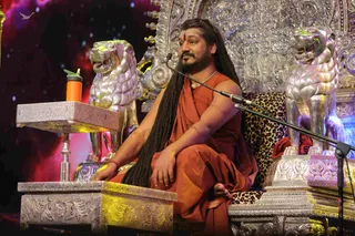 A seated man in an orange-saffron robe with long, swept-back dreadlocked hair and a red tilak on his forehead sits on an ornate silver throne flanked by two silver lion sculptures. A leopard-print cushion covers the seat, and a microphone stand is positioned at his right. A small silver side table holds an orange bottle. The background is a dark starry blue and red backdrop. Several cues here, including the saffron clothing, long hair, ceremonial silver throne and the page's claim, match the appearance-based Swamiji (probable) label, though the face and details are not conclusively confirmed.