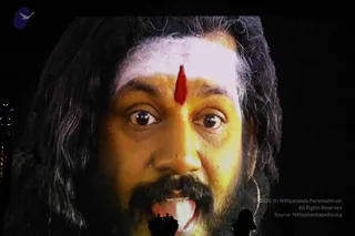 A close-up frame from a video of a man with long, dark, swept-back hair and a full black moustache and beard, looking directly toward the camera with wide eyes and his tongue slightly out. A vertical red mark. The background is black with a faint ornate golden edge at the left. Faint silhouettes of a crown and a small figure appear near the bottom edge. A copyright line and source credit appear at the lower right. Identity is unsure because the face and appearance are not distinctive enough to confirm the page's claim.