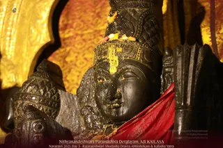 Close view of a dark stone deity murti in a temple shrine, with a carved face, a tall ornate crown, and a yellow turmeric or kumkum mark on the forehead. Yellow powder and small pieces of fruit or flowers sit on the crown, and the face is wet with oil or abhishekam liquid. A red cloth drapes over the shoulder against a backdrop of gold-embroidered fabric. No physical people are visible. The caption at the bottom reads Nithyanandeshwara Paramashiva Devalayam, Adi KAILASA, Navaratri 2021, Day 3, Rajarajeshwari Shodasha Dravya Abhishekam & Kalasha Yatra.