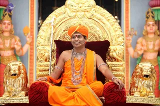 A seated man in orange-saffron robes sits cross-legged on an ornate golden throne with a deep red velvet cushion and golden lion armrests. He wears an orange turban with a red bindi, layered white beaded necklaces, and white beaded armlets, and smiles while a small microphone is clipped near his mouth. Flanking the throne are two large painted deity statues with golden crowns and jewelry, one holding a fan-like object and the other holding a trident-like staff. A white ceremonial fly-whisk stands beside the throne. Swamiji (probable): the orange robes, orange turban and layered beaded necklaces are consistent with the page's identification of Swamiji as present, though appearance alone is uncertain.