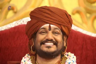 Close-up photograph of a smiling man with a full dark beard wearing a rust-orange turban with a small red tilak mark on his forehead. He wears large gold hoop earrings and a garland of white and pink flowers around his neck, with gold chains visible at the collar. He sits against a deep red upholstered backrest with gilded yellow ornamental carving behind him. Swamiji (probable): the orange turban, ornate gold jewellery and floral garland are the qualifying cues for this person seated on a throne-like chair.