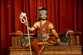 A seated man on an ornately carved wooden throne with leopard-print cushions, set before deep red drapes. He wears a leopard-print headband topped by a silver crescent, saffron-orange cloth draped over his shoulder and body, a leopard-print waist cloth, layered rudraksha malas and a pendant, gold necklace and earrings. He holds a white serpent-shaped staff in his left hand, with his right arm resting on the armrest and a foot bare. a person, seated on the peetha as the central figure, identified by the crescent, rudraksha and throne cues.