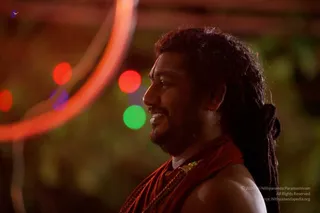 A photograph of a smiling man in profile against a blurred night background with bokeh lights in red, green and purple, and a glowing orange circular light at left. He has long dark hair pulled back with a braid or rope at the back, a short beard and moustache, and wears a reddish-orange shawl draped over bare shoulders with a gold-toned ceremonial necklace.
