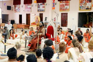 A photograph inside a hall with pale walls and framed pictures, showing a bearded man with long dark hair seated on an ornate silver-and-red throne. He is bare-chested in an orange-red wrap and wears a rudraksha mala, holding a tall silver staff, and a trident stands beside the throne. Dozens of seated devotees surround him on a mat: many women wear white and orange garments with rudraksha malas, and a few men wear orange. A gilded seat with a deity murti stands in the background, along with a tripod camera, a standing fan and a side table with a small object. Hmm, the scene reads as a darshan or satsang.
