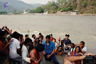 A photograph of a group bathing in a fast-flowing, muddy-brown river, with forested hills and a riverside building behind. In the centre, a man in a blue polo shirt and dark trousers, wearing glasses and a dark rudraksha mala, stands waist-deep in the water among devotees. A bald elderly man in an orange-brown garment and a rudraksha mala stands nearby, and a bare-chested man in saffron-orange cloth is being held and bathed by others. Several women in white and orange clothes are wading, some with hands raised to their heads, and a trident is planted in the water on the left. Onlookers on the bank wear white, orange and red clothing. The and the source line are printed over the lower right corner