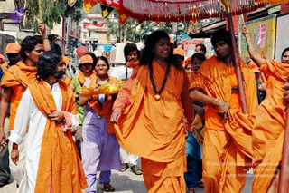 Outdoor street procession in a town, with a red and gold canopy with tassels overhead. A long-haired man in an orange saffron robe and dark rudraksha mala with a gold pendant walks centrally, gesturing with his hand. Beside him a man in saffron wrap and white dhoti walks, and a woman in a striped lavender-and-white salwar holds a brass tray. Other devotees in orange shawls and orange caps follow, and a man on the right in orange robes holds a canopy pole. Shops and signs line the street behind them.