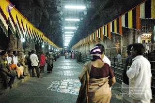 A dimly lit temple corridor with a floral garland-draped ceiling line in yellow, orange, white and purple flowers, and rows of fluorescent tube lights overhead. Painted kolam designs in white and coloured patterns decorate the stone floor. On the left, several uniformed men in khaki and a few in white shirts and orange dhotis sit on a raised stone bench. Devotees walk along the aisle; a woman in a red sari and a woman in a gold-bordered sari stand near the centre. On the right, a woman in a maroon blouse and striped headscarf faces away toward a metal railing and a brightly lit shrine beyond. A man in a white shirt stands at the right edge.