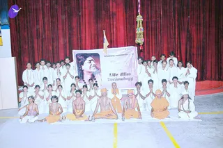 A group photograph in an indoor hall with red drapes behind. A large banner reads "Nithyananda Meditation Academy" and "Life Bliss Technology" beside a portrait of a smiling figure with long dark hair. Many people in white clothing sit and stand in rows; several kneel in front with hands pressed together in anjali. Seated in front, a few younger boys wear orange cloths, rudraksha malas and sacred threads, and one seated figure in orange with a shaved head sits centre-front beside a brass kalasha-style pot and lamps on a white sheet. A yellow floor line marks the stage edge, and a small flag and a tassel decoration hang above.