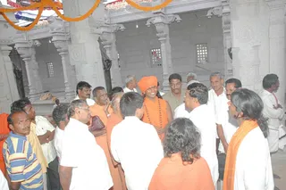 A photograph inside a grey stone temple hall with carved pillars and latticed windows, decorated with orange marigold garlands along the top. A crowd of men in white dhotis and shirts, along with several men in orange and saffron clothing, gathers in a tight circle. A man at the centre wears a saffron turban and an orange shawl with a rudraksha-style beaded necklace, smiling as he speaks with those around him. Another person in an orange shawl stands at the lower right with long dark hair. Figures in white shirts and striped clothing surround the group.