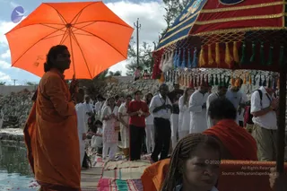 Outdoor ceremony beside a stone-edged water tank. In the left foreground, a man in an orange robe holds a large orange umbrella with a visible emblem and stands near the water's edge. Behind him, a line of standing devotees in white and red clothes press their palms together in prayer on a striped cloth mat. A red and gold canopy with blue and green tassels stands at right, and a woman with braided hair and an orange-robed figure are in the lower right foreground. Some people wear white kurtas and shirts, and one wears a grey shirt with a backpack.