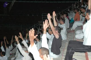 A photograph of a large seated audience in an open-air or tiered auditorium at night. Many people in white ritual clothing sit on the steps with their arms raised high, palms open, in a gesture of devotion or attentive participation. A few wear dark or orange garments. Metal railings and stairs run along the upper left, and rows of empty seats extend into the background. No individual is clearly identifiable as the speaker; no person is confidently recognisable as a person in this crowd, so the presence of a person cannot be confirmed from appearance.