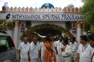 A photograph of an ornate white and terracotta entrance gate with a crenellated top, gold-capped columns and a flower garland arch over the passage, bearing a blue banner reading Nithyananda Spurna Programme. A portrait of a man in white head cloth is placed above the gate. In the center, a long-haired man in an orange saffron robe with a flower garland walks forward, with several men in white shirts and white dhotis with rudraksha malas around them, one man with palms joined in greeting, and a silver vehicle at left. Hanging decorative lanterns hang on either side of the gate. Shown are about fifteen people in total.