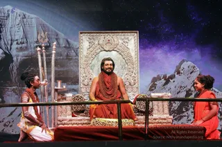 A stage set at an event with a seated man in orange-red robes and a long rudraksha mala with a large bead necklace, sitting on a silver and jewelled throne with leopard-print cushions. Two attendants sit on the stage floor: one on the left in white dhoti with a yellow sash, a rudraksha mala and a braided topknot, the other on the right in an orange sari with a rudraksha mala. Silver trident-topped staffs and a silver kalasha stand to the left of the throne. The backdrop shows a snowy mountain and a starry sky. A railing separates the stage from the viewer. The seated man has the appearance of a person, with long swept hair, orange robes and rudraksha malas, seated on the throne as the central figure.