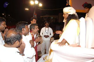 A night-time outdoor event on a raised stage with bright floodlights overhead. A man with long dark hair, wearing a white turban, white robe and a yellow-gold shawl, sits on a white throne-like seat with white cushions and gestures toward a group. Several men in white shirts and white dhotis, one wearing a red-and-gold shawl, sit or stand with palms pressed together in respectful greeting. A younger man in a white shirt and beige trousers wears a rudraksha mala and stands with hands folded. Beyond the stage, rows of red chairs and a white-lit building are visible. Swamiji appearance is a probable match: long swept-back hair, white turban and gold-trimmed shawl on a throne.