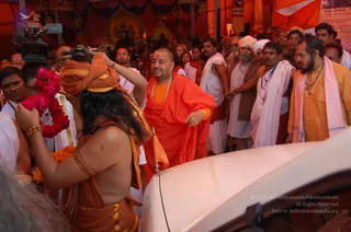 A crowded outdoor evening scene under an orange tent canopy with a deity image in the background. In the foreground, a figure with long dark hair, wearing an orange wrap and gold-toned bangles, holds a pink flower garland and turns toward a man in a saffron-orange robe with a rudraksha-style beaded necklace and a red tilak on his forehead, who gestures with one hand near a white car. Around them, men in white dhotis and shawls, a man in a white turban, and a man in an orange kurta with a pink shawl and beaded necklace press close. A camera operator on the left films the scene, and a cameraman with a video camera stands in the upper left. Orange flags and a printed banner hang behind the crowd.
