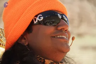 Close-up photograph of a smiling man wearing a bright orange knit beanie and reflective sunglasses with the letters DG set in sparkling stones on the temple. His long dark hair falls behind his ear, and a leopard-print shawl is visible at the lower left. A small microphone headset is positioned near his mouth. The reflection in his sunglasses shows a crowd of people gathered on a hillside under a pale sky, and the background is a blurred dry, tan landscape. The page indicates the event but the face and clothing cues do not establish identity, so no identification is made.