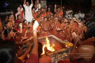 A night-time homa at an outdoor fire pit, with a large square metal fire trough in the foreground where a bright flame rises. Around it, many devotees in saffron-orange robes with rudraksha malas and white tilaka on their foreheads raise their arms and chant. Some are bare-chested in saffron cloths, others wear saffron kurtas. Several women in red and orange saris stand at the left, hands folded or raised. A brass bowl of offerings and small brass vessels sit on the fire pit's edge. A man in a white kurta with a rudraksha mala and a raised arm stands near the top center. Behind them are parked cars, a few onlookers in white, and a lit pillar or altar at the right edge.