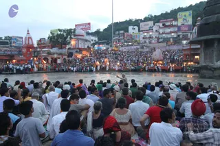 Evening ganga aarti at a riverside ghat with a wide, dense crowd of devotees seated and standing on the steps and in the water's edge, many holding up mobile phones to photograph. A figure in white near the water raises a lit lamp toward the river while others in white stand nearby. Temple spires, painted shrines, illuminated hoardings and a hillside with hotel buildings frame the scene. A small shrine stands on the right near the flowing river.