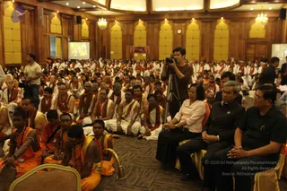 A large wood-panelled hall with yellow cushioned wall panels and chandeliers holds a seated audience of many devotees in white dhotis and white shirts with red shawls, plus several in saffron-orange robes with rudraksha malas and bangles in the foreground. Three guests sit in front-row chairs on the right: a woman in a pale pink blouse and dark skirt, and two men in dark formal clothes. A cameraman on a tripod films from the centre aisle, and projection screens on both sides show images. The scene suggests a seated satsang gathering.