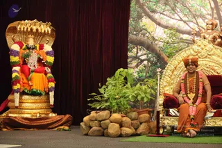 A seated man in orange and saffron robes sits on an ornate gold throne with red cushions, wearing a saffron turban, a rudraksha mala, a gold pendant necklace and a pink and white flower garland, with bangles on his wrists. A deity murti in red and orange robes sits on a gold throne under a golden multi-hooded serpent canopy, draped in colourful flower garlands of purple, yellow, red and white. Behind them a painted backdrop shows a large banyan tree, and a bronze statue of a figure stands on the upper right. Potted plants and stacked rocks sit in front of the stage, with a green carpet and maroon velvet curtains. Swamiji (probable) seated on the throne, a candidate by saffron-orange robes, turban, rudraksha mala and ceremonial gold necklace.