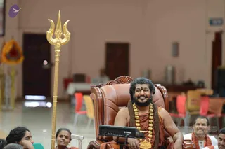 A man seated in a brown leather throne-style chair smiles broadly during a satsang hall gathering. He wears a rust-orange sleeveless garment, a rudraksha mala, long heavy gold necklaces, and a headset microphone, with long swept-back dark hair and a short beard. A tall golden trident stands to the left. Seated devotees in the foreground include women and a man in white with an orange-trimmed shawl. Behind them are red and yellow chairs, a white cloth-covered table, and a wall with a framed picture and a doorway. A Yamaha keyboard or mixer sits on a small table in front of the throne.