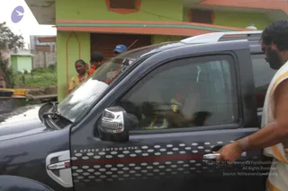 A dark grey SUV with a 5 Speed Automatic decal and white polka-dot side graphics is parked on a street. A man in a white dhoti with an orange-and-gold border stands at the open front door on the right, one hand on the handle. Through the tinted rear window, a figure in ornamented clothing is partly visible inside. Behind the vehicle, a woman in a yellow sari and a child stand near a green-and-orange house with a rolling shutter. A small blue cap is visible among the onlookers.