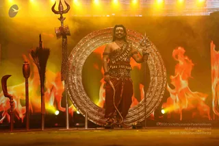 A man stands on a stage in front of a large ornate golden circular frame, smiling, with a trident held in his right hand and a curved ceremonial sword and other staffs arranged to the left. He wears a leopard-print wrap over one shoulder, a leopard-print belt, a red-brown dhoti, rudraksha-style bead bracelets and a necklace, with a red-brown tilak on his forehead. Behind him stand glowing orange flame backdrops under warm yellow stage lights, and a small wall of fire imagery surrounds the set. Stage props include a tall trident-topped staff with a cloth banner and a feathered staff. The scene reads as a theatrical stage presentation rather than a religious ceremony.
