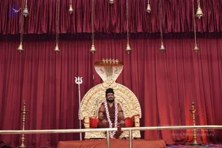 A seated man in saffron-red robes sits cross-legged on an ornate gold throne with a carved cobra-hood backrest and red cushions, centred on a stage before deep red draped curtains. Hanging from the top edge are brass bells on cords. He wears a white and pink flower garland, a rudraksha mala, and a horizontal white forehead mark, and his hair is tied up. A brass trident stands to the left of the throne. Brass oil lamps stand on either side of the platform, and a gold railing runs across the front.