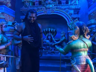 A photograph taken inside a temple-style display lit in blue. A smiling man with long dark hair and a large black rudraksha mala draped over his chest and shoulders stands with his right hand on his chest, wearing a dark sleeveless black garment and beaded wrist bands. Behind him are painted life-size statues of deity figures in green and orange tones, with a multi-headed serpent canopy above a shrine niche holding a deity murti decorated with a garland. A female figure is depicted in the shrine area.