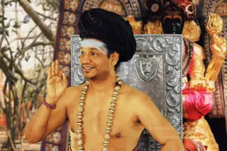 A bare-chested man with a tall black turban and a white horizontal forehead mark stands in front of an ornate silver-embossed backrest decorated with a lion face. He wears a long necklace of large gold beads and a red thread bracelet on his left wrist, and raises his right hand in a blessing gesture while smiling. Behind him stands a gilded deity murti with a raised golden arm and a pink garland, set among an ornamental golden frame and a blurred outdoor background of bare trees.