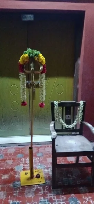 A brass trident (trishul) stands upright on a yellow square base on a red and grey patterned tiled floor, its prongs decorated with yellow and red marigold garlands, green leaves and strings of white jasmine hanging down. Beside it is a dark wooden chair with a white jasmine garland draped over its backrest. Behind them is a green-painted door set in a red wall. No people are visible.